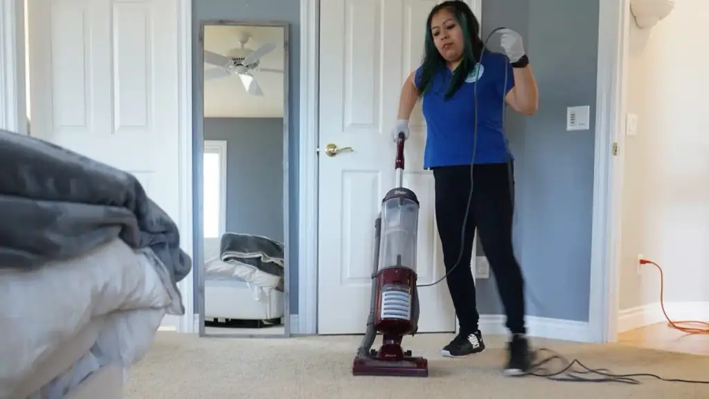 A cleaner in a blue logo polo vacuuming a bedroom carpet with an upright vacuum