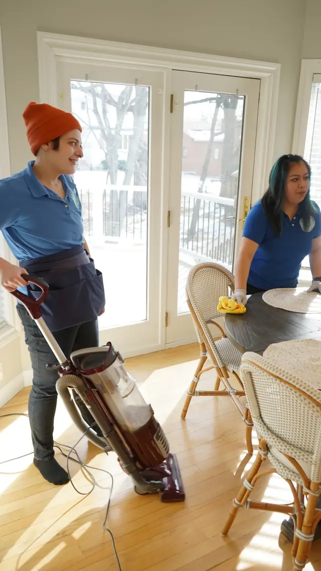 Two cleaners finishing a sunlit dining room, one vacuuming, one wiping the table